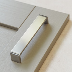 May include: A brushed nickel cabinet pull with a rectangular shape and rounded edges.