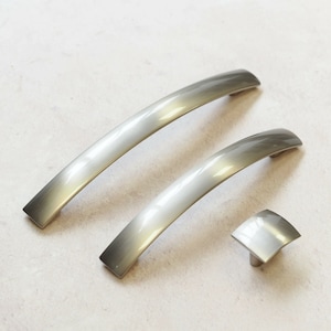 May include: Three brushed nickel cabinet pulls, two are curved and one is square. The pulls have a gradient finish, going from a darker nickel to a lighter nickel.
