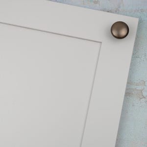 Peut inclure: Gros plan sur une porte d'armoire blanche avec un bouton de couleur bronze dans le coin sup&eacute;rieur droit. La porte pr&eacute;sente un design &agrave; panneaux encastr&eacute;s. Le bouton rond a une finition m&eacute;tallique.