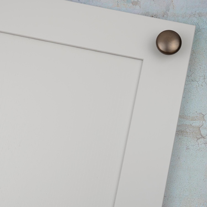 Peut inclure: Gros plan sur une porte d'armoire blanche avec un bouton de couleur bronze dans le coin sup&eacute;rieur droit. La porte pr&eacute;sente un design &agrave; panneaux encastr&eacute;s. Le bouton rond a une finition m&eacute;tallique.