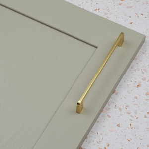 Könnte beinhalten: Ein goldfarbener Schrankgriff ist an einer hellgrauen Schranktür montiert. Der Griff ist rechteckig und hat ein schlichtes, modernes Design. Die Schranktür hat eine eingelassene Platte und eine strukturierte Oberfläche.
