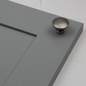 Könnte beinhalten: Ein silberfarbener Schrank-Knopf mit einer runden, leicht gewölbten Oberseite. Der Knopf ist an einer grauen Schranktür montiert.