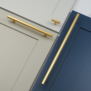 May include: Gold-coloured cabinet handles and a T-knob are shown on grey and navy blue cabinet doors. The hardware has a modern, minimalist design. The cabinet doors have a wood grain texture. The long handle is approximately 20 cm long.