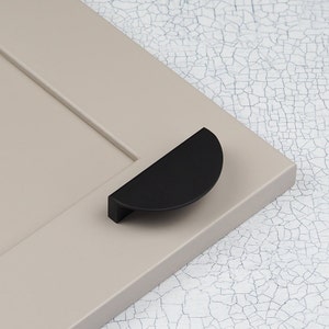 Może przedstawiać: Matowy czarny uchwyt do szafki w kształcie półksiężyca jest zamontowany na jasnobeżowych drzwiach szafki. Uchwyt ma nowoczesny design, o prostej, czystej estetyce. Drzwi mają wpuszczany panel.