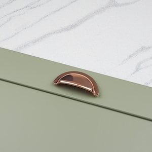May include: A copper cabinet knob with a rounded, arched shape. The knob is mounted on a sage green cabinet door.