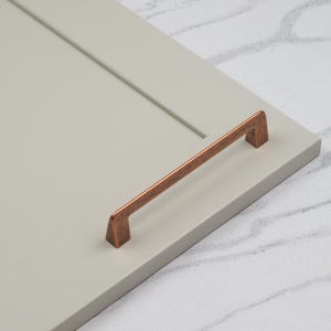 Könnte beinhalten: Nahaufnahme einer kupfernen Schranktürgriffe mit gebürsteter Oberfläche. Der Griff ist an einer hellgrauen Schranktür montiert.