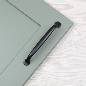 May include: A black cabinet pull handle mounted on a light green cabinet door. The handle is a curved bar with rounded ends. The door has a recessed panel design.