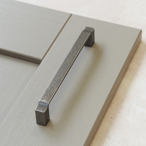 Puede incluir: Un tirador de armario de metal gris oscuro con forma rectangular. El tirador está montado en una puerta de armario gris claro.