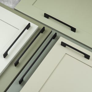 May include: Four matte black cabinet pulls with a linear, rectangular design are mounted onto light sage green and off-white cabinet doors. Each handle features a slim, horizontal bar with two perpendicular mounting posts at the ends.