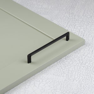 Peut inclure: Poignée de porte de placard noire de forme rectangulaire avec des bords arrondis. La poignée est montée sur une porte de placard vert clair.