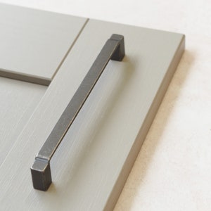 May include: A black metal cabinet door handle with a rectangular shape. The handle is mounted on a light gray cabinet door.