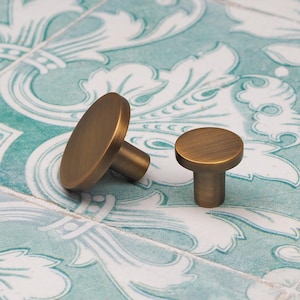 May include: Two antique brass cabinet knobs. One knob is oval-shaped, and the other is round. The knobs are on a patterned, light blue and white surface. These knobs are ideal for adding a vintage touch to furniture.