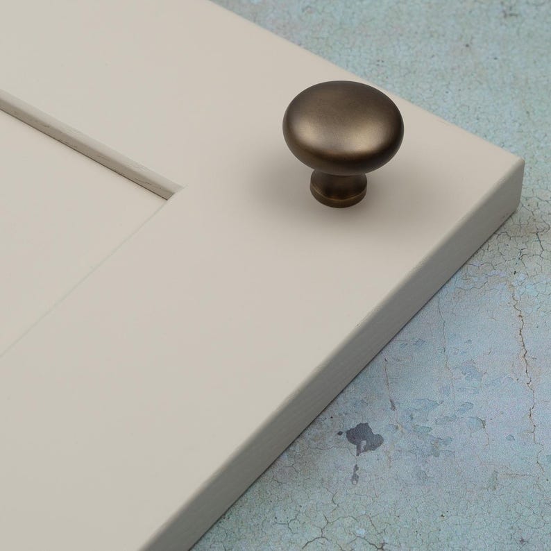 Peut inclure: Gros plan sur une porte d'armoire cr&egrave;me avec un bouton de couleur bronze. Le bouton est rond et poss&egrave;de une courte tige. La porte de l'armoire est rectangulaire et poss&egrave;de un panneau encastr&eacute;.