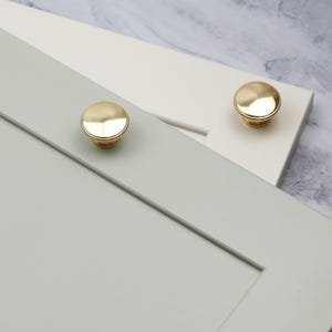 May include: Two gold cabinet knobs with a round, slightly raised design. The knobs are shown mounted on cabinet doors, one on a light green door and one on a white door.