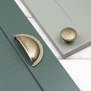 May include: Close-up of two cabinet door samples, one in a deep green and the other in a light grey. A bronze-coloured cup pull and a round knob are displayed on the doors, showcasing hardware options for kitchen or bathroom cabinetry.