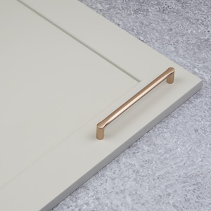May include: A brushed rose gold cabinet pull with rounded edges. The pull is mounted on a white cabinet door.