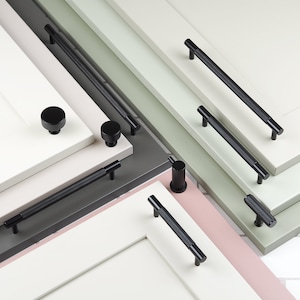 Puede incluir: Surtido de pomos y tiradores de armario negros en varios tamaños y estilos. Los pomos y tiradores se muestran en superficies de diferentes colores, incluyendo blanco, verde y rosa.