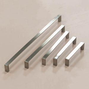 May include: Four silver cabinet pulls of varying lengths, arranged in a triangular pattern on a light pink surface.