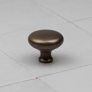 Peut inclure: Un bouton de meuble en bronze avec un dessus arrondi et une base courte et cylindrique. Le bouton est pos&eacute; sur une surface carrel&eacute;e blanche. Ce mat&eacute;riel convient aux tiroirs, aux armoires et autres meubles.