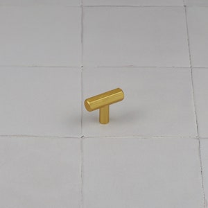 Pode incluir: Uma maçaneta de armário em forma de T de cor dourada com acabamento escovado.