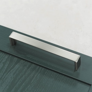 Może przedstawiać: Rektangularny, zaokrąglony uchwyt do szafki wykonany z szczotkowanego niklu. Uchwyt jest zamontowany na ciemnozielonych drzwiach szafki.