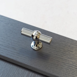 Pode incluir: Uma maçaneta de armário cromada com uma pega redonda e uma barra reta. A maçaneta está montada numa superfície cinzenta escura.