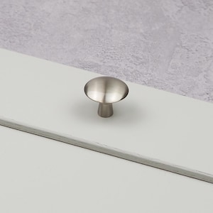 May include: A brushed nickel cabinet knob with a mushroom-shaped design. The knob has a rounded top and a short, cylindrical base. The hardware is set against a white surface and a textured gray background.