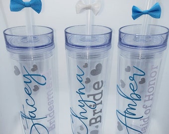 May include: Three clear plastic tumblers with straws and personalized text. The tumblers are decorated with silver glitter hearts and blue and white bows. The text on the tumblers reads "Stacey Bridesmaid", "Shryna Bride", and "Amber Maid of Honor".