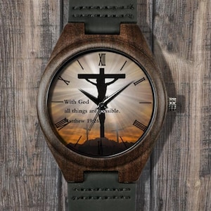 May include: A wooden wristwatch with a dark brown case and a green leather strap. The watch face features a silhouette of a cross with the text "With God all things are possible. Matthew 19:26."