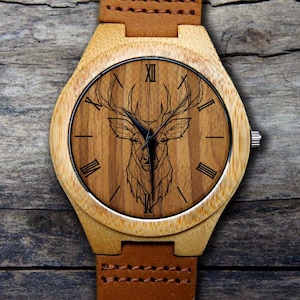 May include: A wooden wristwatch with a brown leather band. The watch face features a geometric deer design with Roman numeral hour markers.