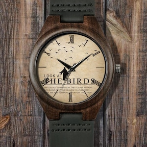 May include: Wooden wristwatch with a brown case and a cream-colored face. The watch face features black Roman numerals, black hands, and a silhouette of a person with birds. The text "LOOK AT THE BIRDS" is printed on the watch face.