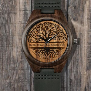 May include: A wooden wristwatch with a dark brown case and a light brown face. The watch face features a tree design with the text "Jesus said to them, I am the bread of life" and "John 6:35". The watch has a dark green leather strap.