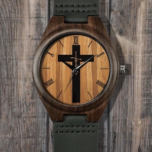 Puede incluir: Reloj de pulsera de madera con esfera de madera marrón con diseño de cruz negra. El reloj tiene una correa de cuero verde oscuro y un bisel de madera marrón. Los números romanos marcan las horas. El reloj se muestra sobre una superficie de madera.