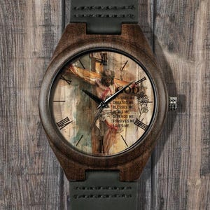 May include: A wooden wristwatch with a brown case and a dark green leather strap. The watch face features an image of Jesus on the cross with the word "GOD" and the text "DESIGNED ME, CREATED ME, BLESSES ME, HEALS ME, DEPENDS ME, FORGIVES ME, LOVES ME."
