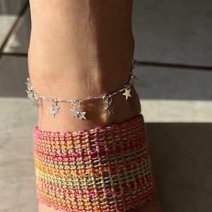 May include: Silver ankle bracelet with star charms. The bracelet is on a person's ankle, and the person is wearing a colorful woven sandal.