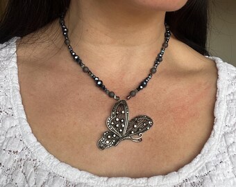 Puede incluir: Un collar de plata con un colgante de mariposa y cuentas negras y grises. La mariposa tiene un diseño detallado con pequeños cristales.