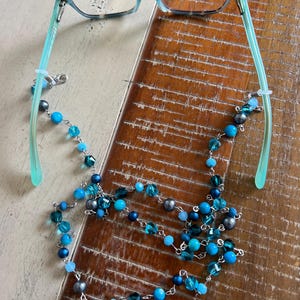 May include: A pair of eyeglasses with light blue and tortoiseshell frames attached to a beaded chain. The chain features a variety of round and faceted beads in shades of turquoise, dark blue, and metallic gray, connected by silver-toned metal links.