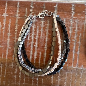May include: A multi-strand bracelet featuring faceted beads in silver, black, and bronze tones. The bracelet has a silver-toned clasp and is displayed on a wooden surface. The bracelet is a jewelry accessory.