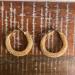 May include: A pair of gold-colored hoop earrings. The earrings have a woven texture and a hinged closure. The hoops are displayed on a distressed, brown wooden surface.