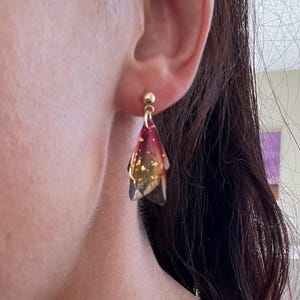 May include: Gold-colored stud earrings with a dangling, translucent, petal-shaped design. The petals transition from a deep red to a golden yellow, with gold flecks throughout. The earrings are a unique and elegant accessory.