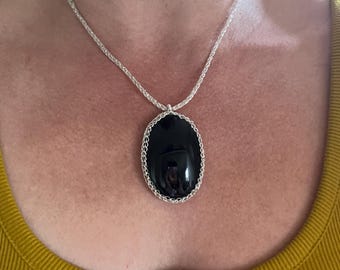 May include: A silver necklace featuring a large, oval black pendant. The pendant is framed by a silver chain design and hangs from a delicate silver chain. The necklace is displayed against a yellow top.