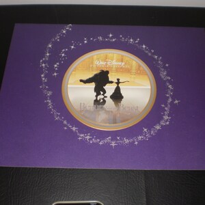 Op de afbeelding: Een paarse ingelijste print met een gouden cirkel met een silhouet van Belle en het Beest dat danst. De cirkel is omgeven door een zilveren glitterontwerp en de tekst "Walt Disney Pictures Presents Beauty and the Beast".