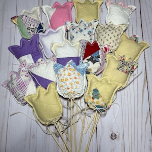 May include: A collection of handmade fabric tulip decorations in various colors and patterns. The tulips are attached to wooden dowels and tied with twine. The fabric designs include floral, plaid, and solid colors, creating a charming, handcrafted aesthetic.