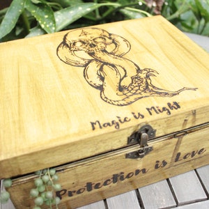Può includere: Una scatola di legno con un coperchio incernierato e una chiusura. La parte superiore della scatola è decorata con un'immagine bruciata a legno del Marchio Oscuro della serie Harry Potter. Il testo "Magic is Might" è anche bruciato a legno sulla parte superiore della scatola. L'interno della scatola ha il testo "Protection is Love" bruciato a legno sul fondo.