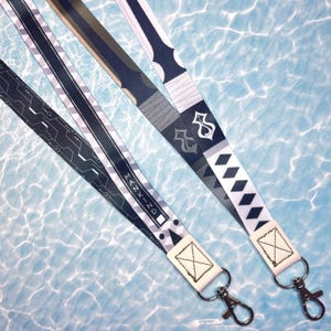 Peut inclure: Deux cordons en tissu arborant des motifs graphiques distincts. L'un présente un design de circuit géométrique sombre avec le texte MANKIND, tandis que l'autre arbore un motif inspiré d'une épée avec un manche blanc, des losanges et un emblème.