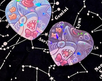 May include: Two heart-shaped stickers featuring cartoon sharks with pink bows, hats, and fish. The stickers have a holographic effect and are set against a black background with a white constellation pattern.