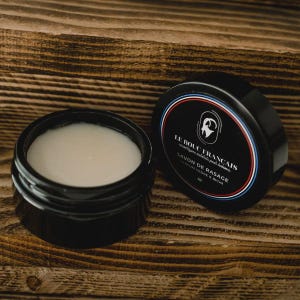 Peut inclure: Deux pots noirs de savon à raser sur une surface en bois. L'un est ouvert, révélant le savon blanc. L'autre est fermé, avec le texte "LE BOUC FRANCAIS" et "SAVON DE RASAGE".
