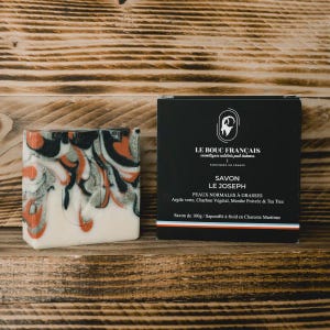 May include: A bar of soap with a marbled design in black, orange, and silver, next to a black box. The box has white text that reads "LE BOUC FRANÇAIS" and "SAVON LE JOSEPH". The soap is for normal to oily skin.
