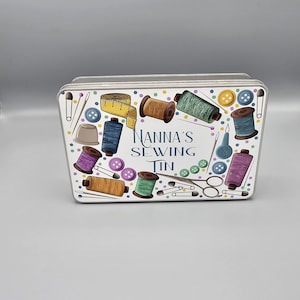 May include: White metal tin featuring a colourful design of sewing supplies, such as spools of thread, buttons, needles, scissors, and a tape measure. The text "Nanna's Sewing Tin" is printed on the tin.