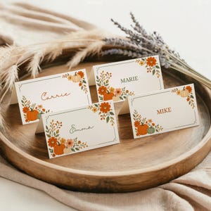 May include: Four white folded place cards featuring a decorative border of orange pumpkins, green leaves, and small flowers. The cards display the names Carrie, Marie, Emma, and Mike in various fonts. Each card has a thin green rectangular frame.